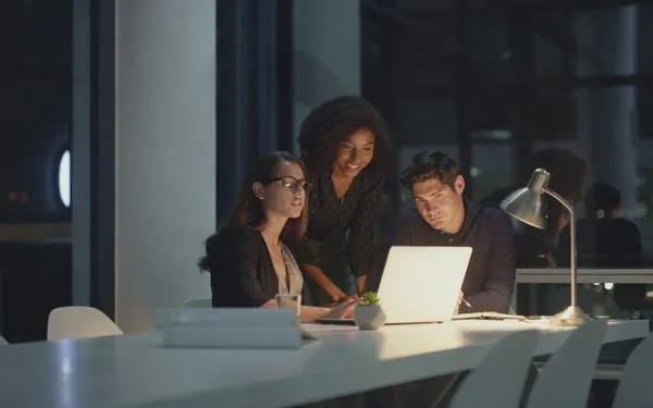 Três profissionais reunidos em volta de um notebook à noite